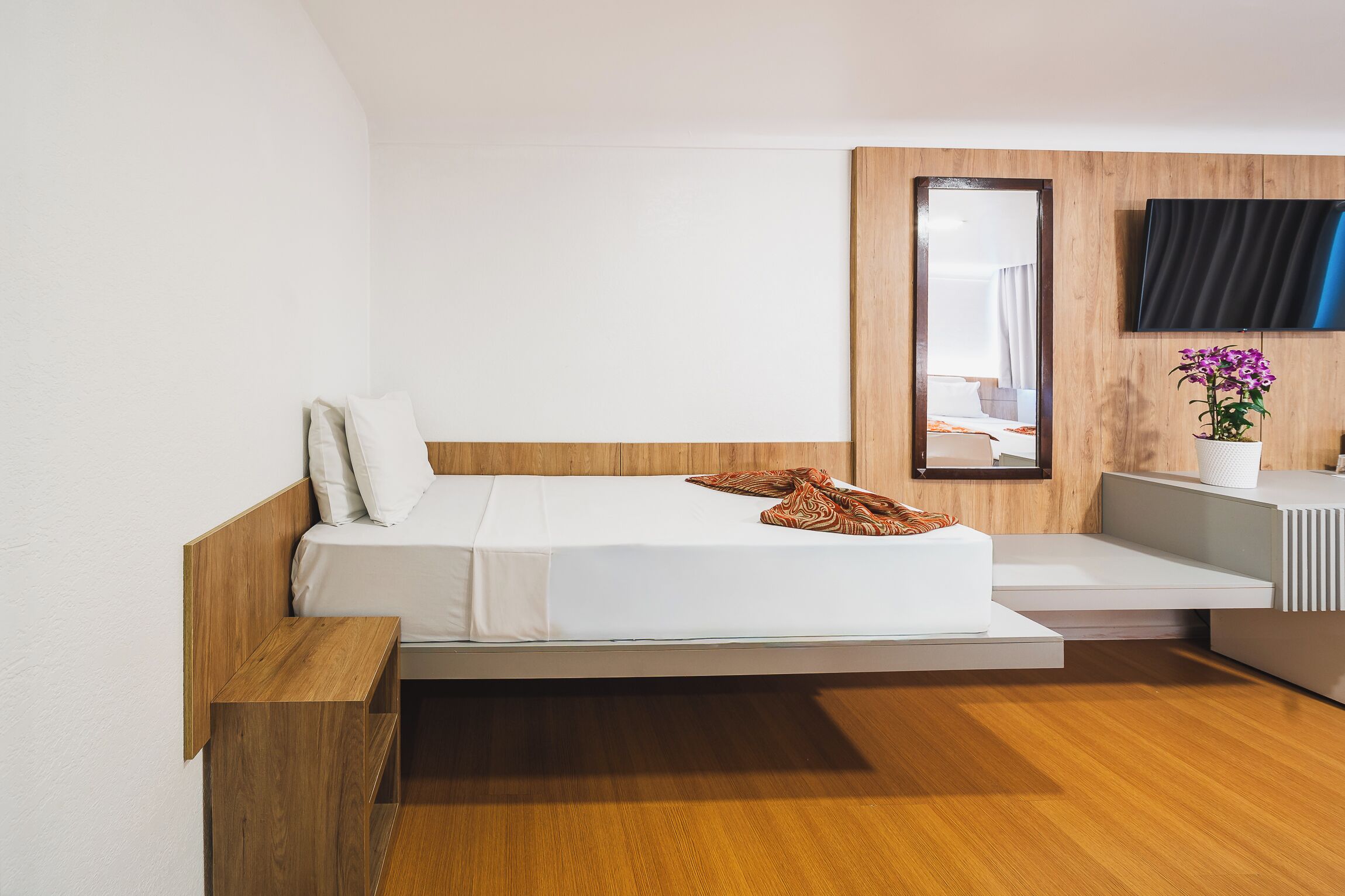 Quarto de hotel moderno com cama, cabeceira de madeira, TV de tela plana na parede e uma pequena planta em vaso.