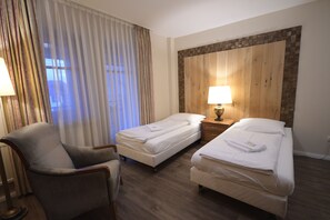 Habitación de hotel con dos camas individuales, una silla, una mesita de noche con lámpara y un ventanal con cortinas.