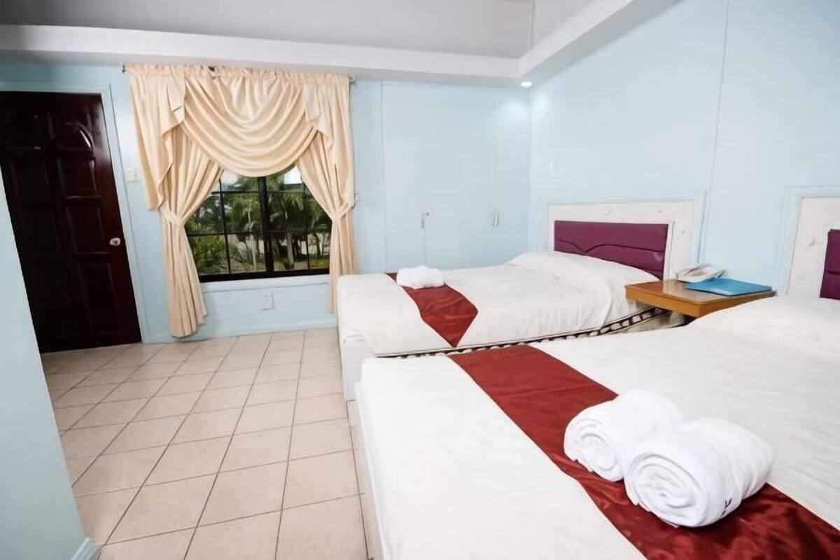 A hotel room with two beds, a window with curtains, a bedside table with a phone, and a view of greenery outside.