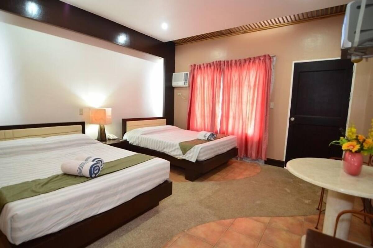 A hotel room with two beds, a small table with a vase of flowers, a door, and an air conditioning unit.