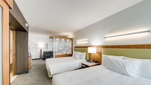 A hotel room with two beds, a wooden headboard, a bedside table with a lamp, a mirror, and a carpeted floor.