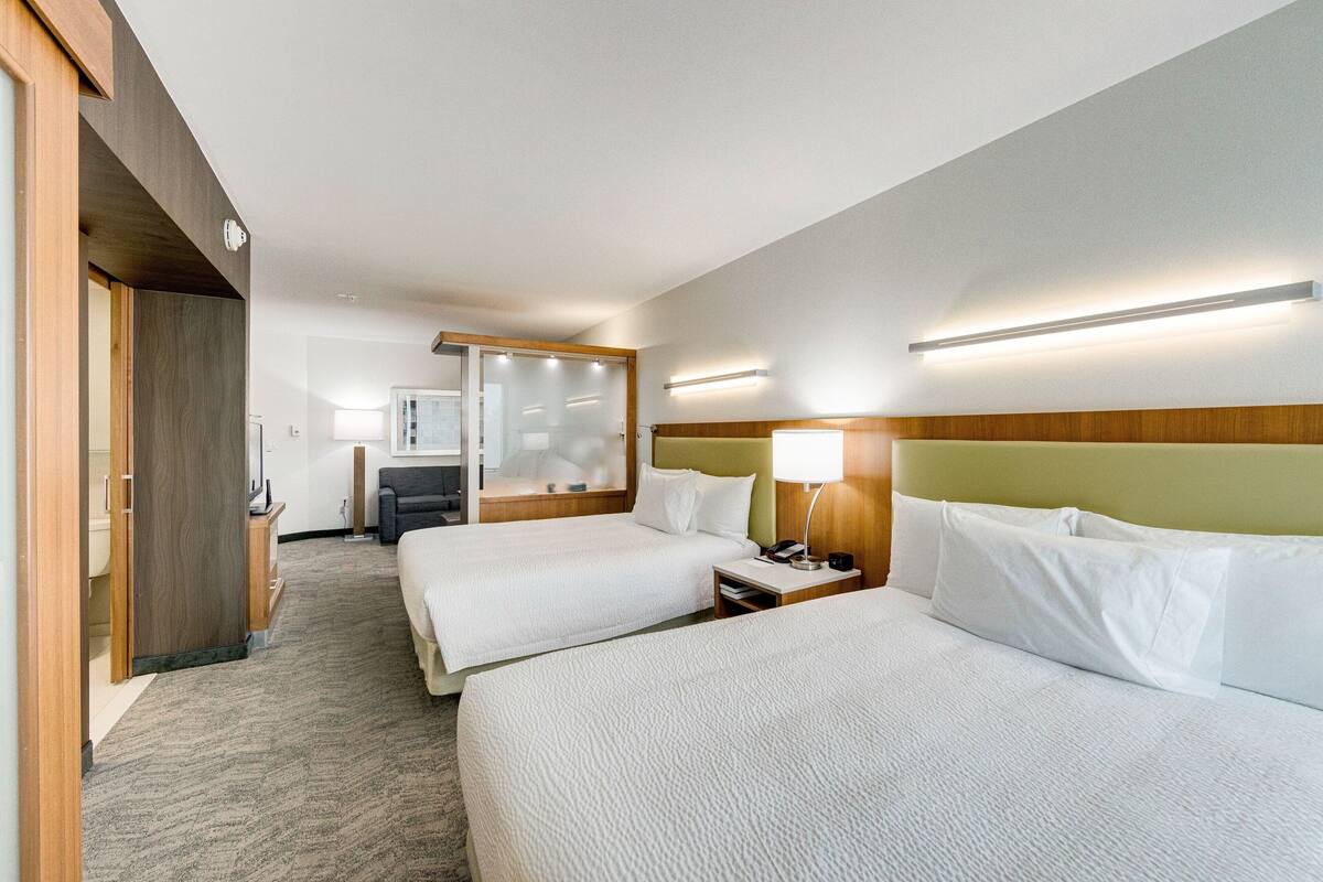 A hotel room with two beds, a wooden headboard, a bedside table with a lamp, a mirror, and a carpeted floor.