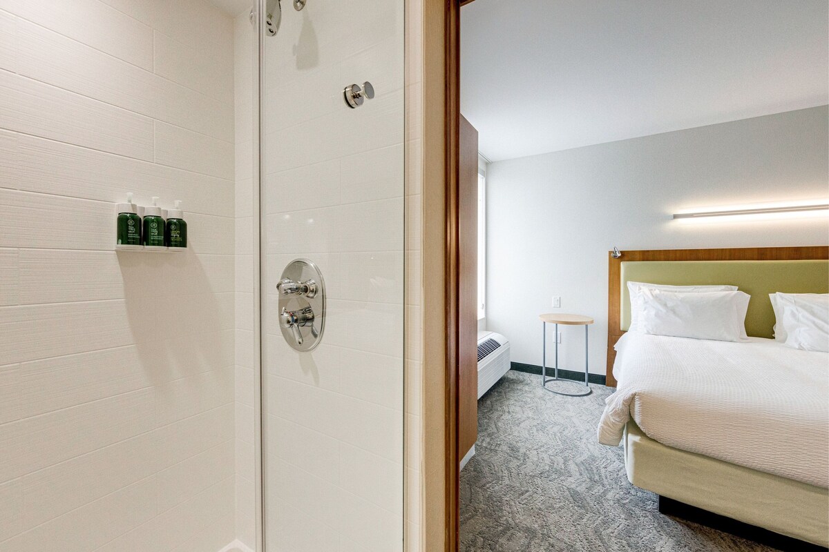 A hotel room with a shower, a bed, a bedside table, and a wall-mounted shelf with toiletries.