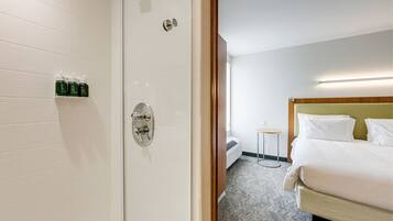 A hotel room with a shower, a bed, a bedside table, and a wall-mounted shelf with toiletries.