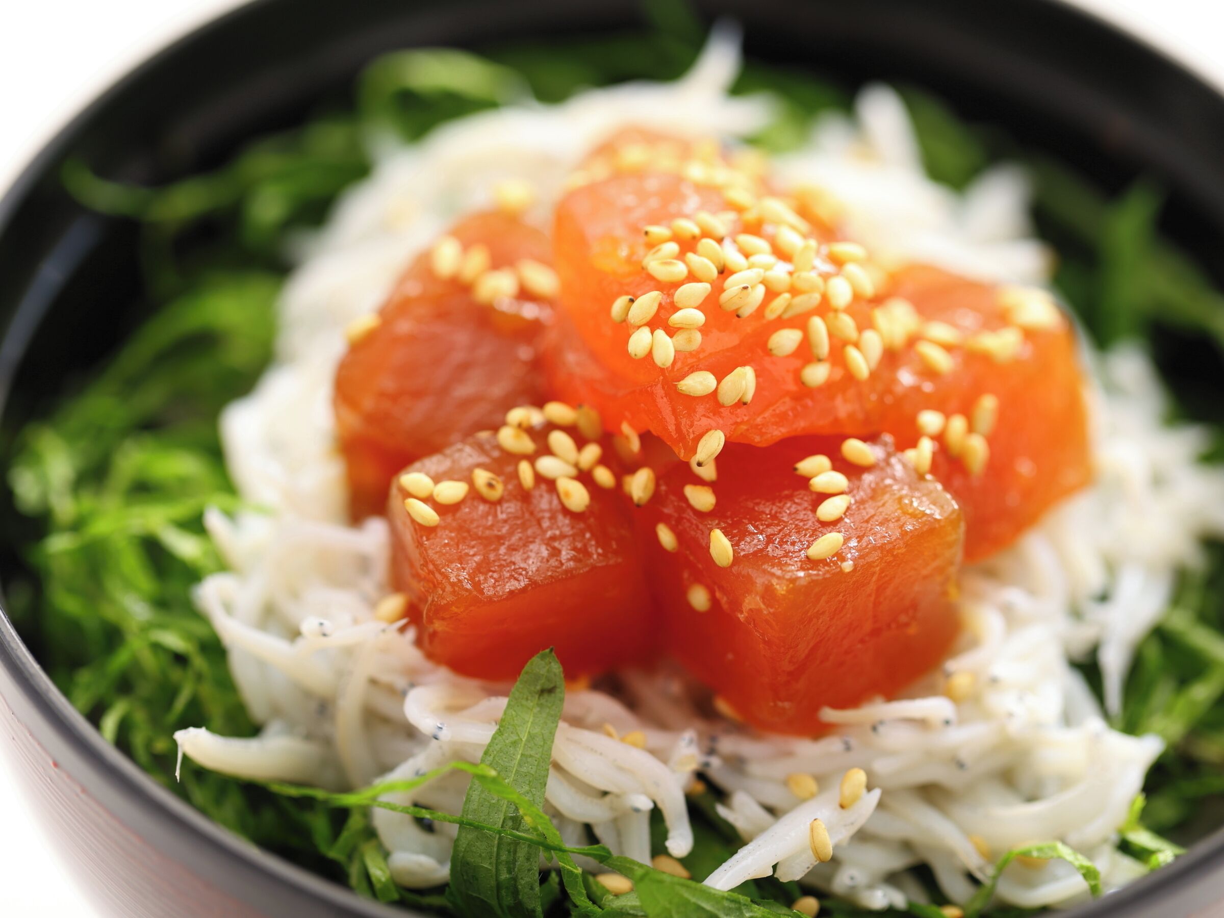 A bowl of food with rice, green leaves, and salmon cubes topped with sesame seeds.