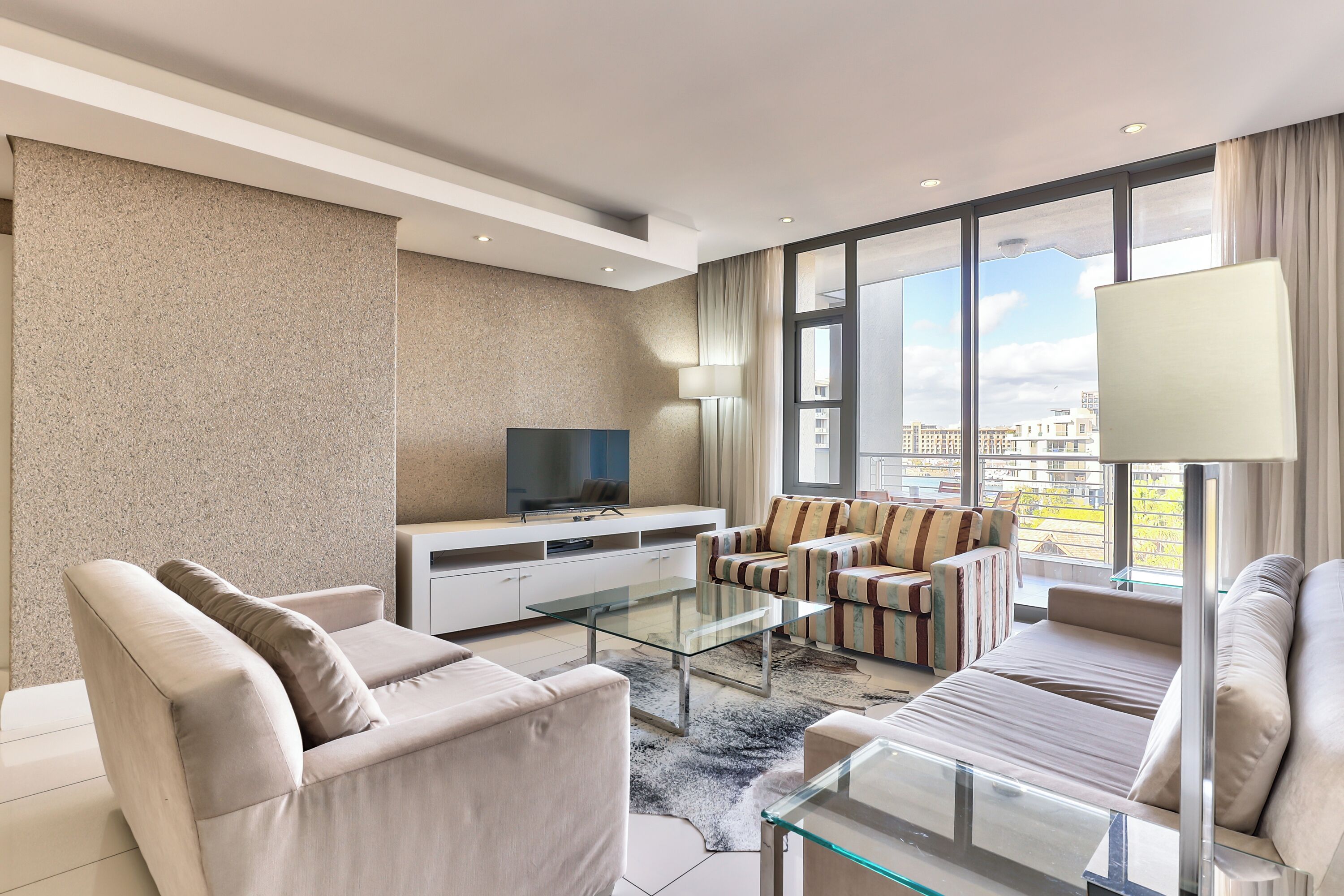 A modern living room with a sofa, armchairs, a glass coffee table, a TV on a white cabinet, and large windows offering a city view.
