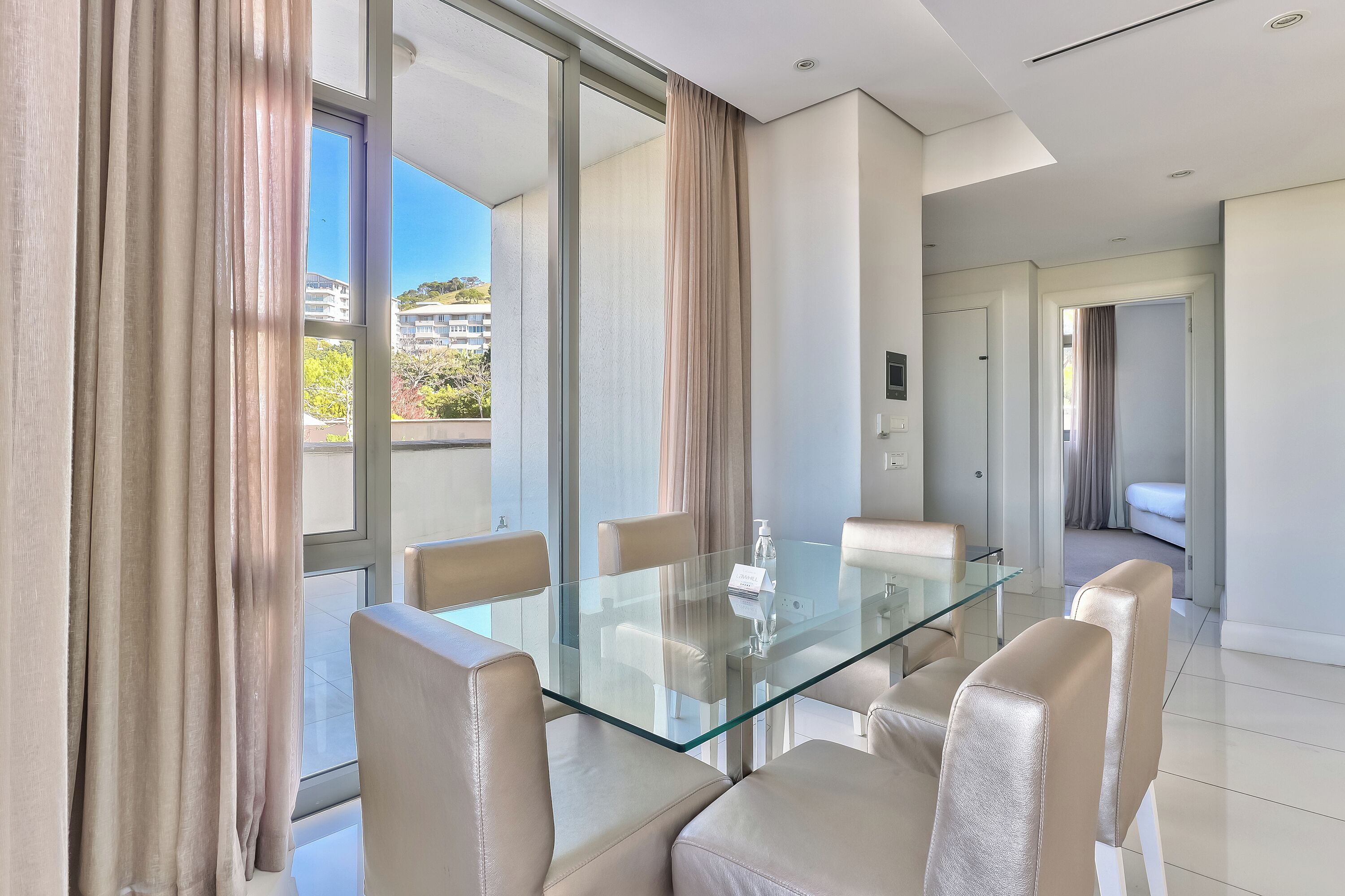 Quarto de hotel moderno com mesa de jantar de vidro, cadeiras bege e grandes janelas com cortinas.