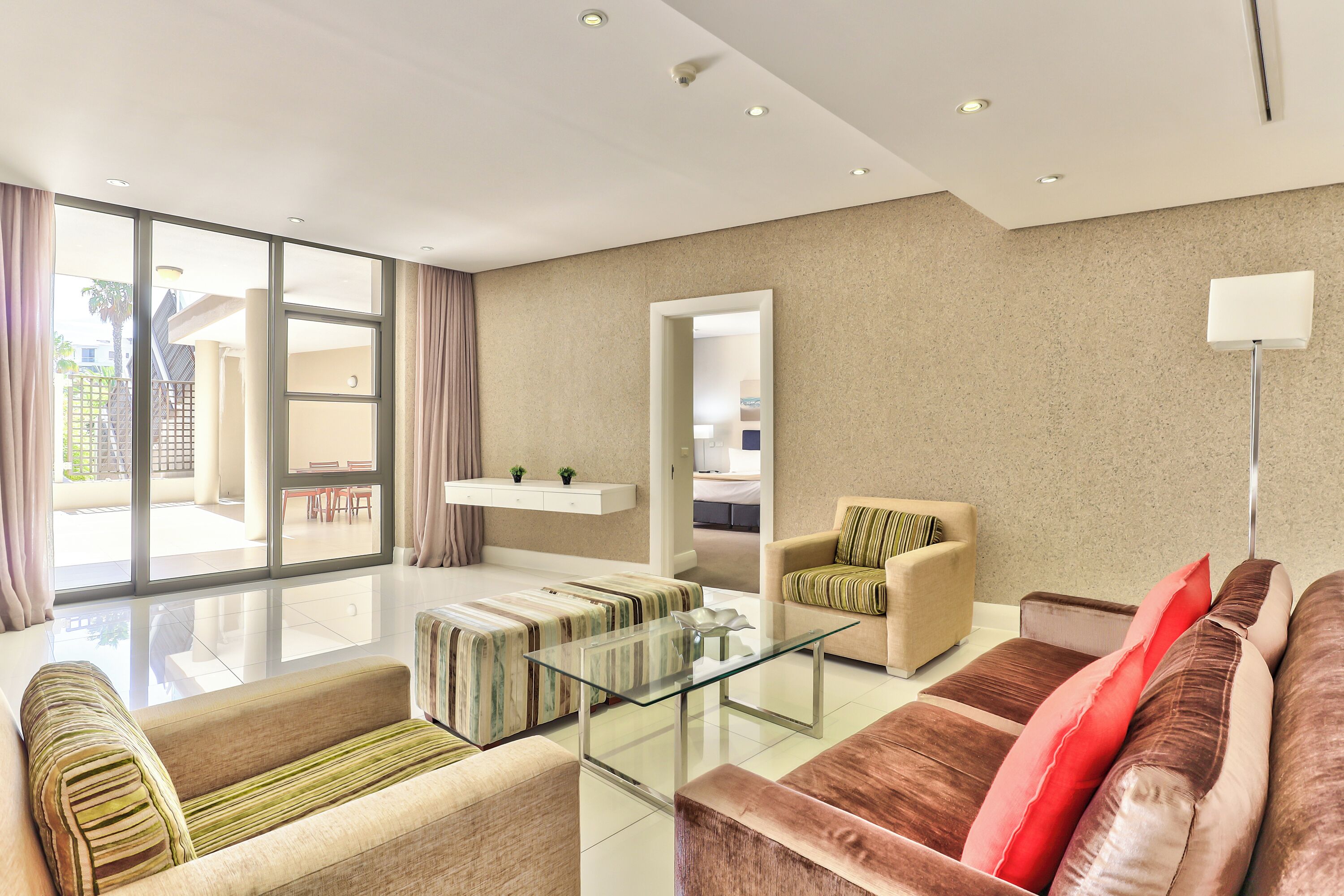 A modern living room with a glass coffee table, sofas, and a large window offering a view of the outdoor area.