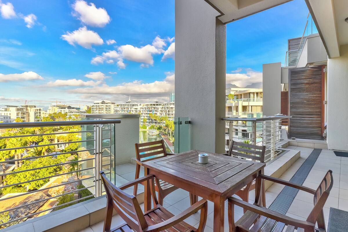 Een balkon met een houten tafel en stoelen, met uitzicht op een woonwijk met moderne gebouwen en groen.