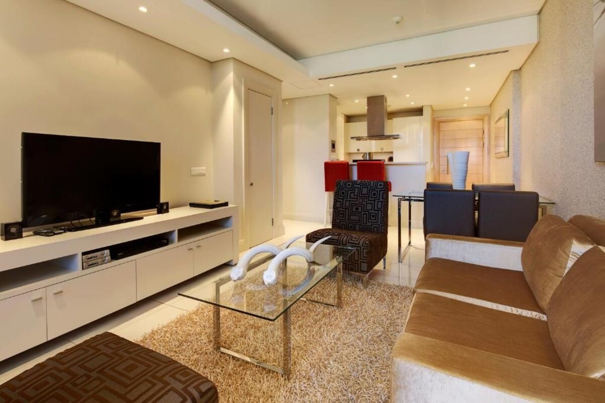 A modern living room with a flat-screen TV, a glass coffee table, and a brown sofa.