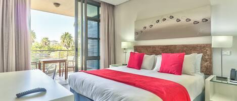 A modern hotel room with a large bed, a desk, and a balcony with a view of palm trees.