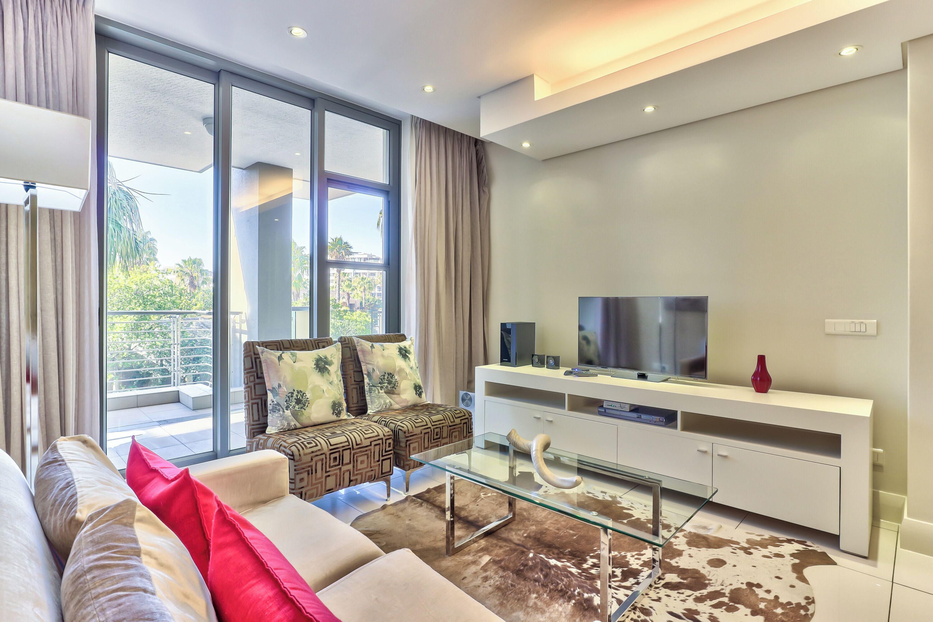 A modern living room with a glass coffee table, a sofa set, a TV on a white entertainment unit, and a balcony with a view of greenery.
