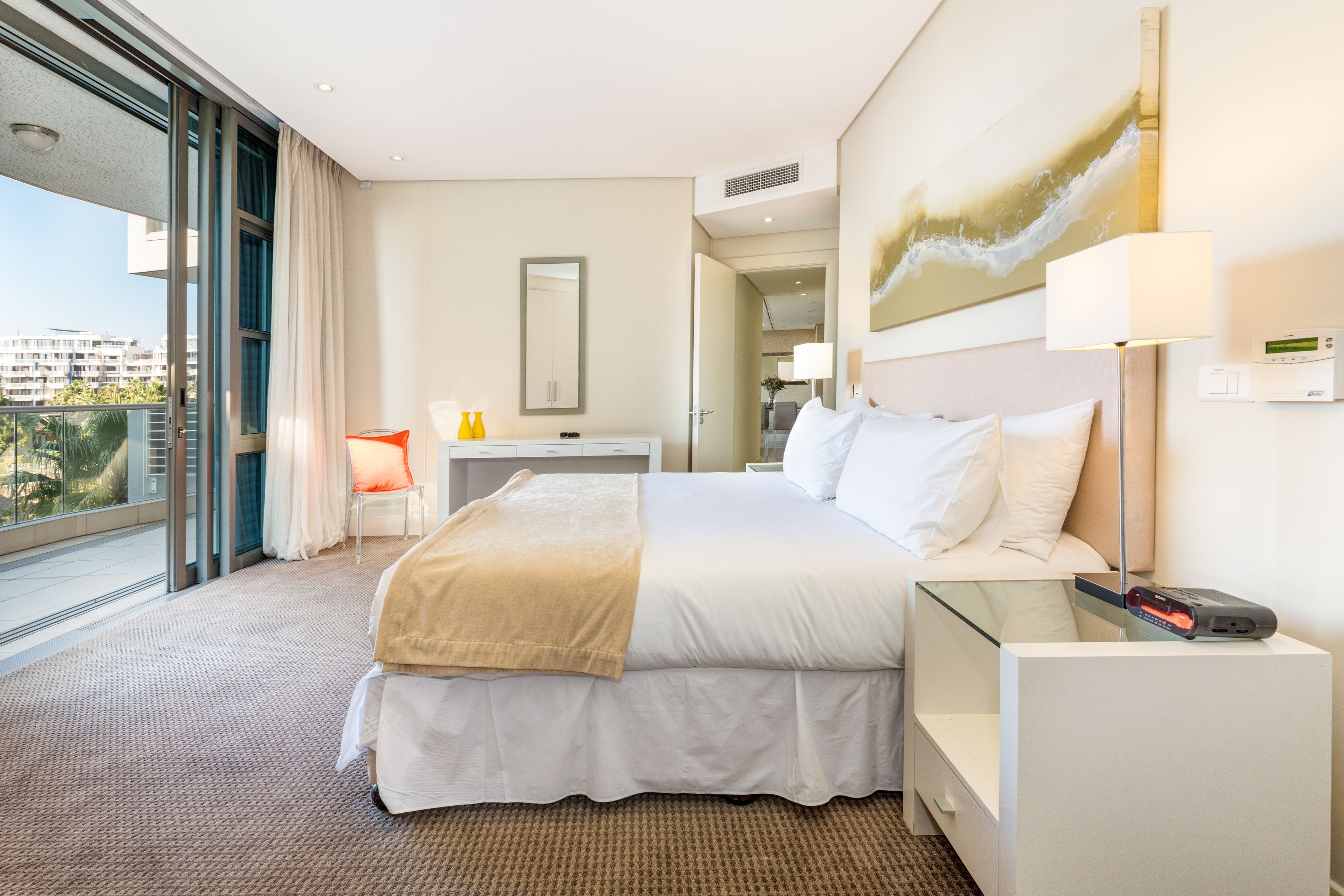 A hotel room with a large bed, a desk, a chair, a glass partition, and a view of the city.