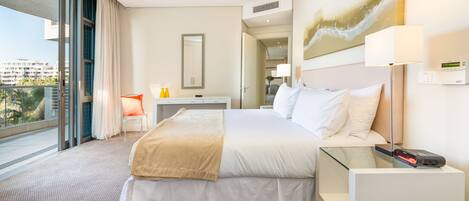 A hotel room with a large bed, a desk, a chair, a glass partition, and a view of the city.