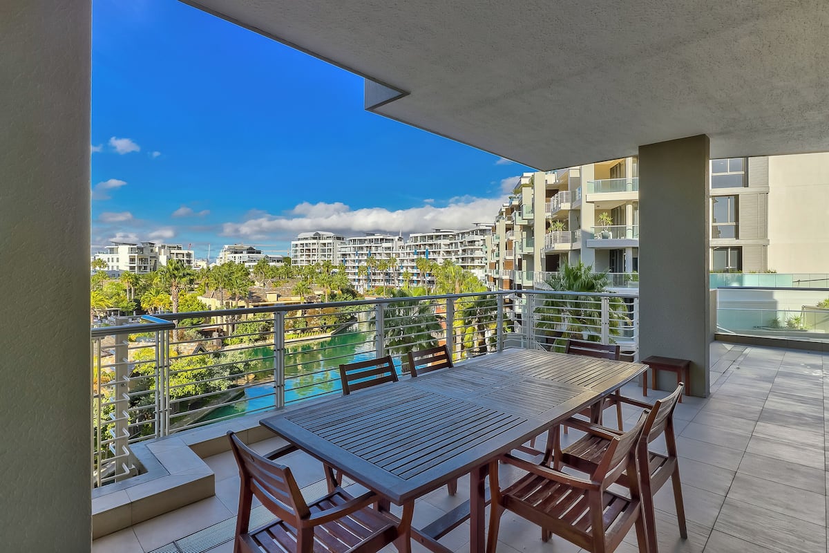 Een balkon met een houten tafel en stoelen, met uitzicht op een zwembad en woongebouwen.
