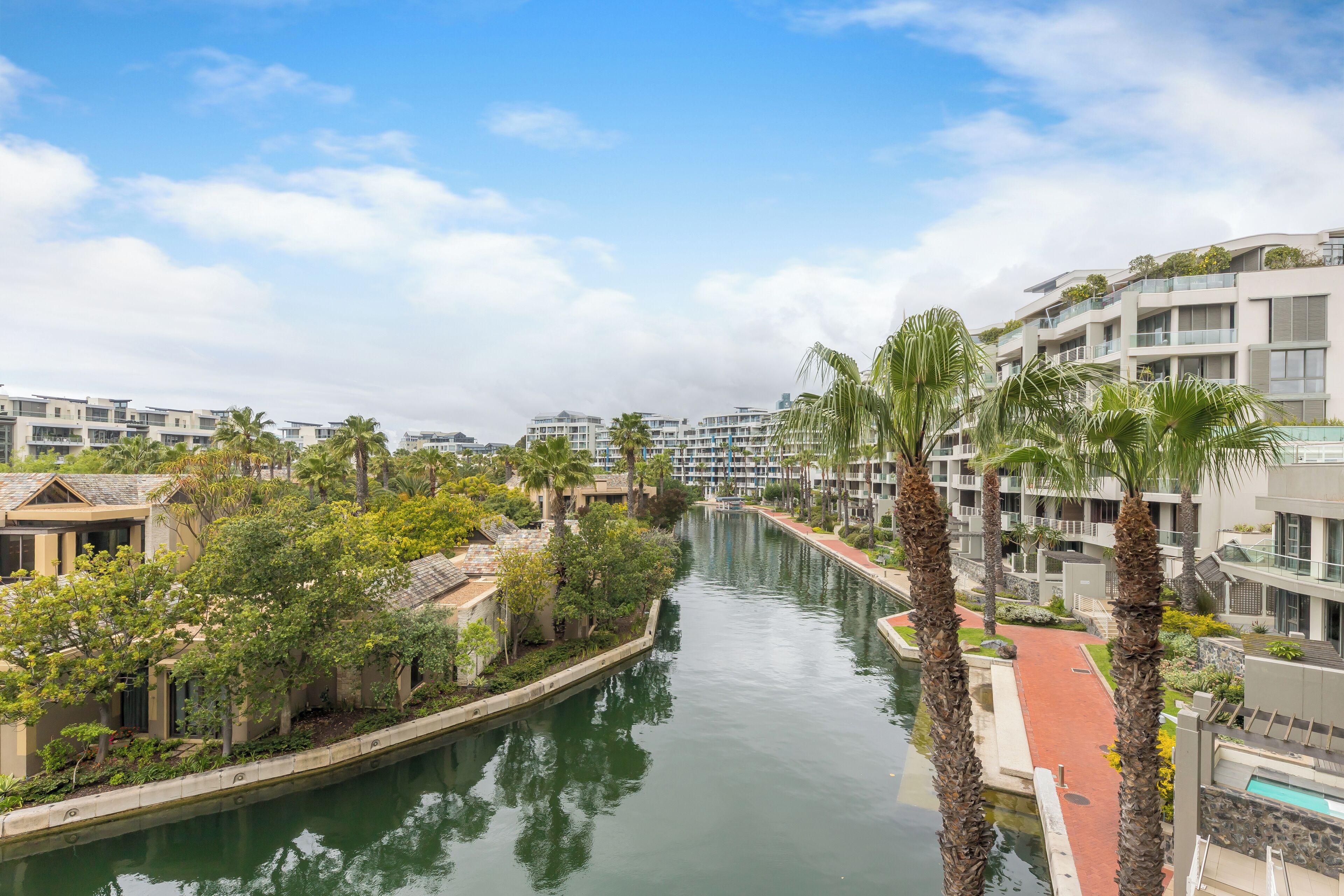 A canal flanked by modern buildings with balconies, surrounded by greenery and palm trees.