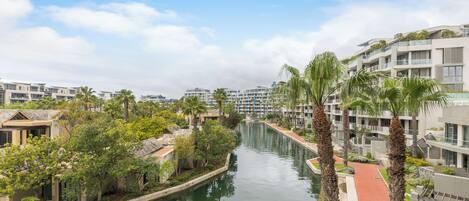 A canal flanked by modern buildings with balconies, surrounded by greenery and palm trees.