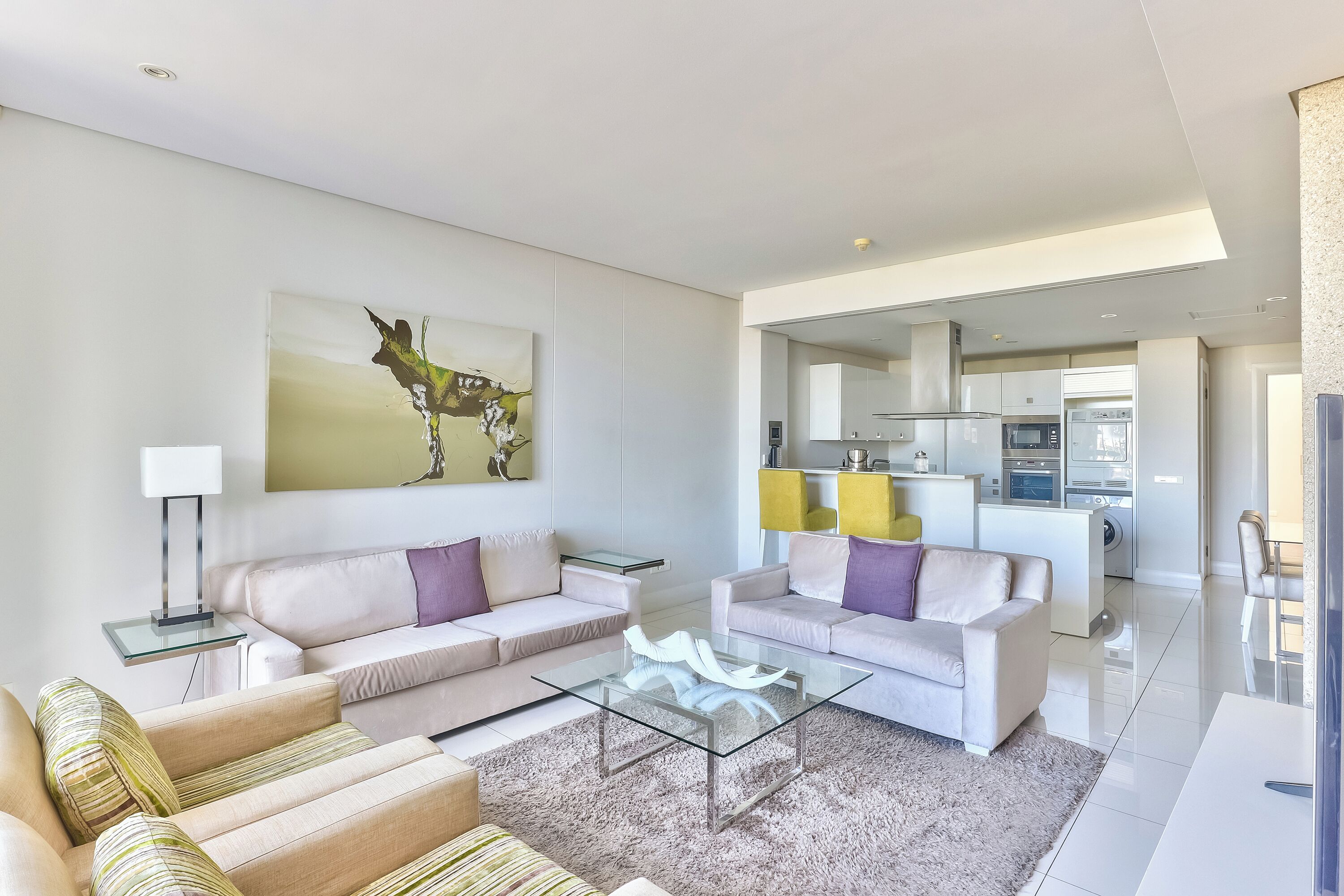 A modern living room with two sofas, a glass coffee table, and a painting on the wall. A kitchen is visible in the background.