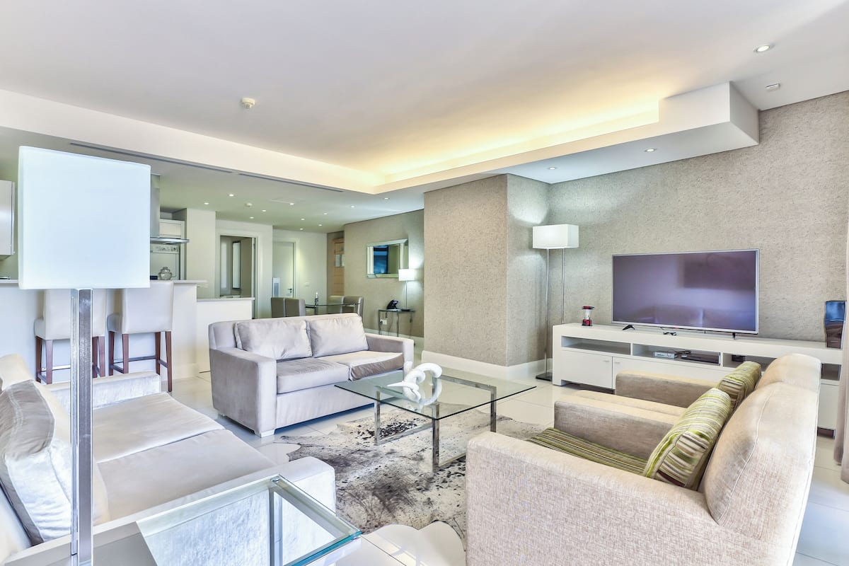Een moderne woonkamer met een bank, fauteuils, een glazen salontafel en een flatscreen televisie.