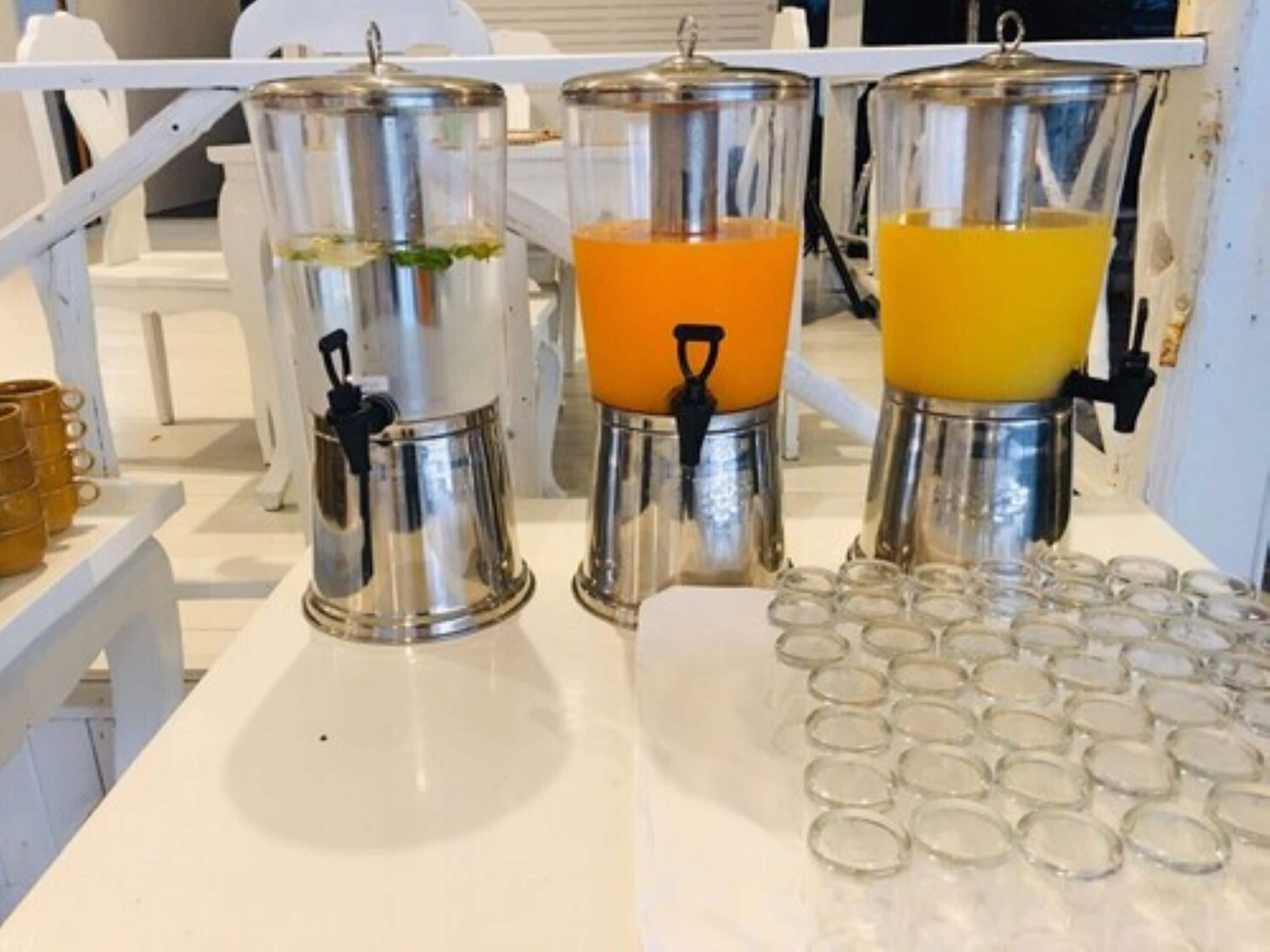 Una estación de bebidas con tres dispensadores, uno con una bebida verde y dos con bebidas anaranjadas y amarillas, y una bandeja de vasos vacíos.