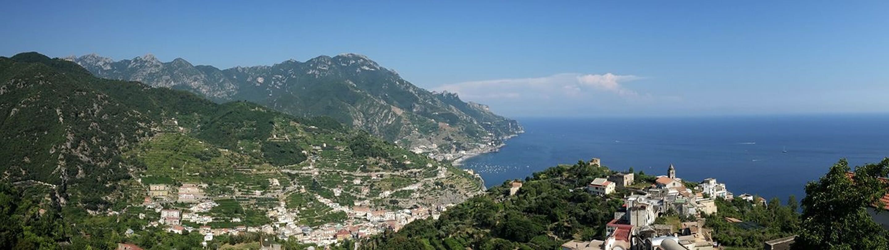 A coastal town with mountainous terrain in the background, a clear view of the sea, and a mix of residential and possibly historical buildings.