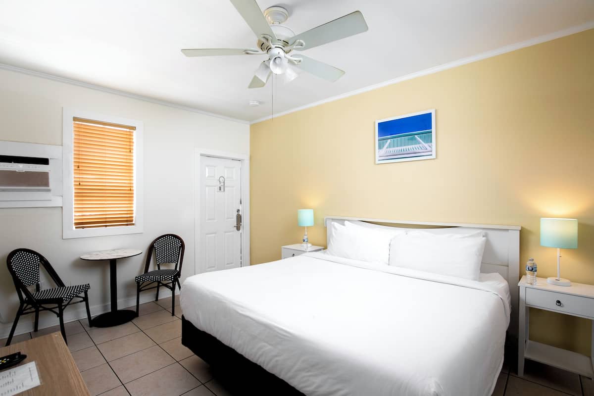 A hotel room with a bed, two chairs, a small table, a ceiling fan, a nightstand with a lamp, and a door.