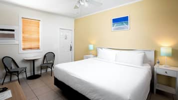 A hotel room with a bed, two chairs, a small table, a ceiling fan, a nightstand with a lamp, and a door.