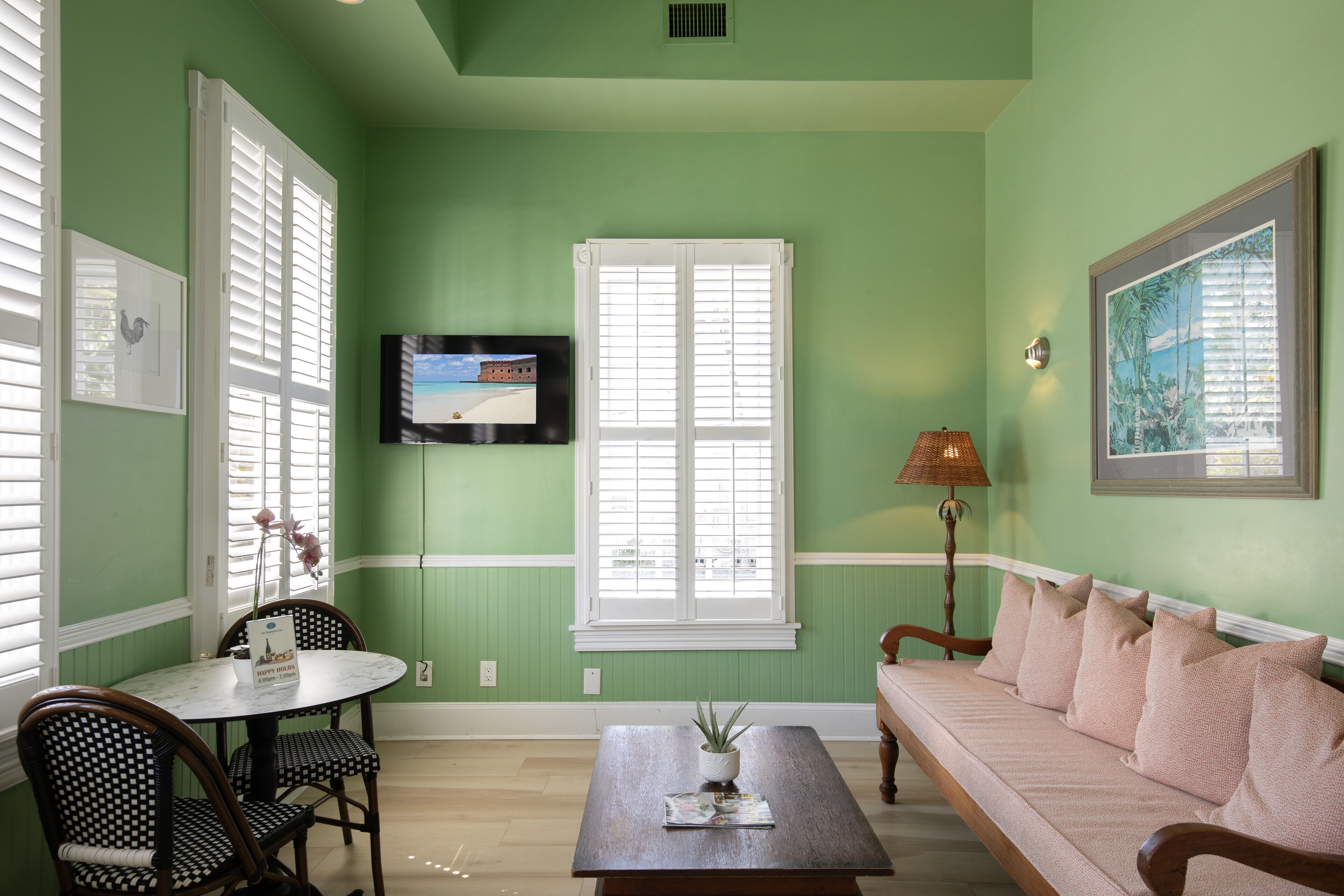 A room with green walls, a round table with chairs, a sofa, a TV mounted on the wall, and a window with shutters.
