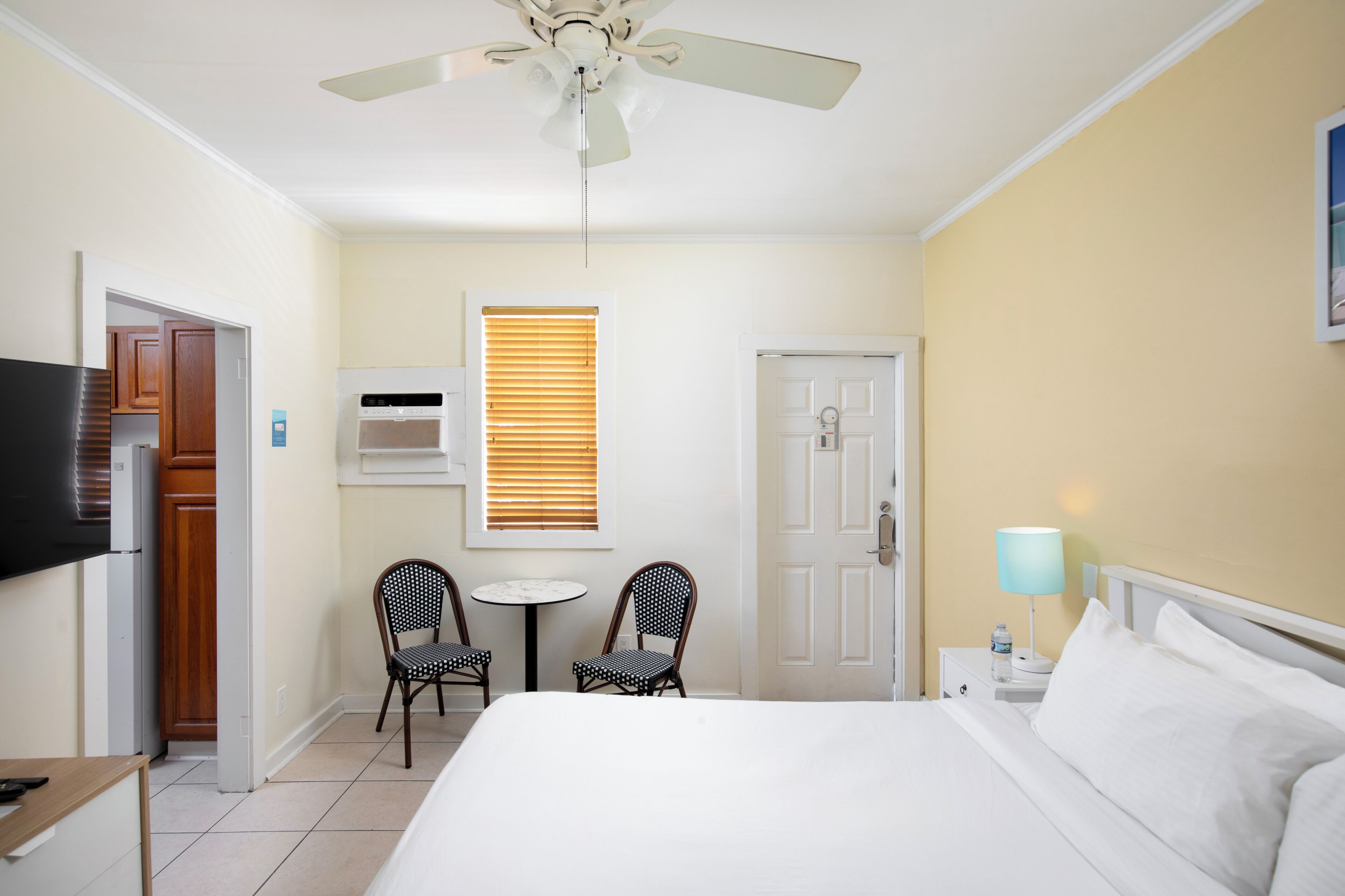 Quarto de hotel com cama, ventoinha no teto, televisão, mesinha com duas cadeiras, janela com persianas e mesa de cabeceira com abajur.
