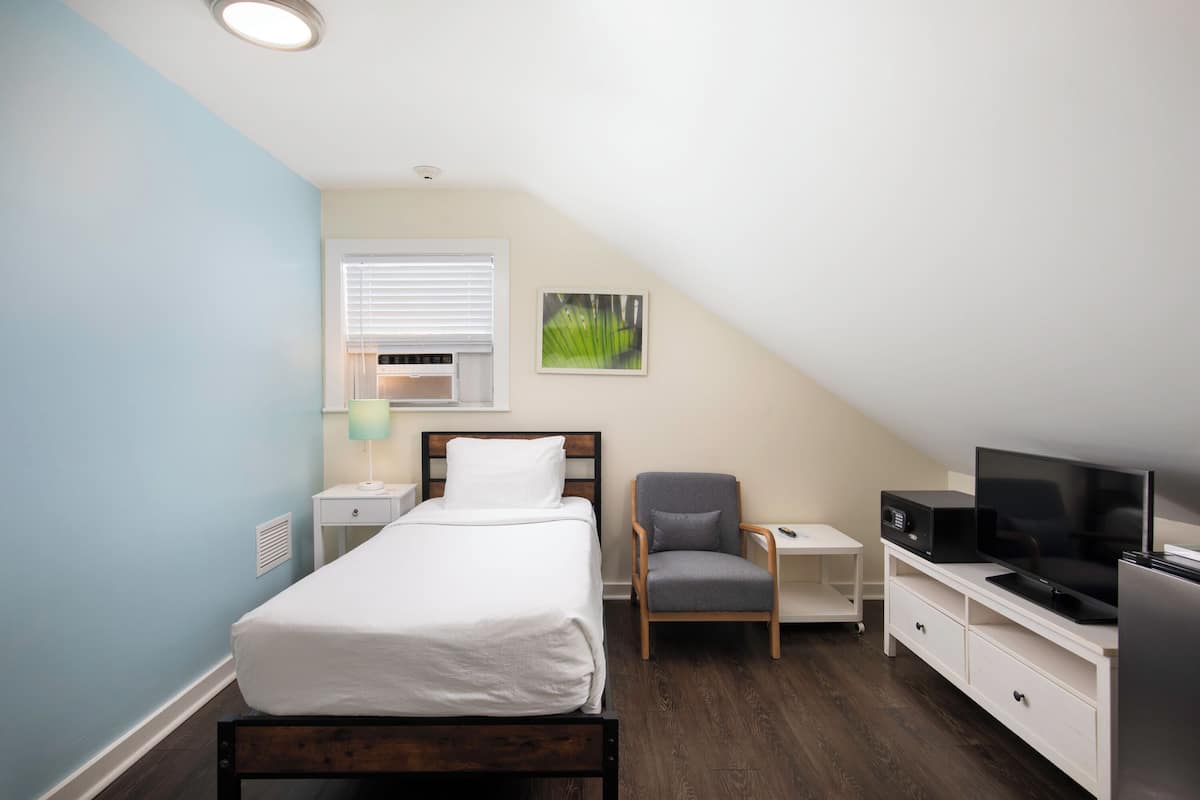 A bedroom with a bed, a chair, a desk with a TV, and a window with blinds.