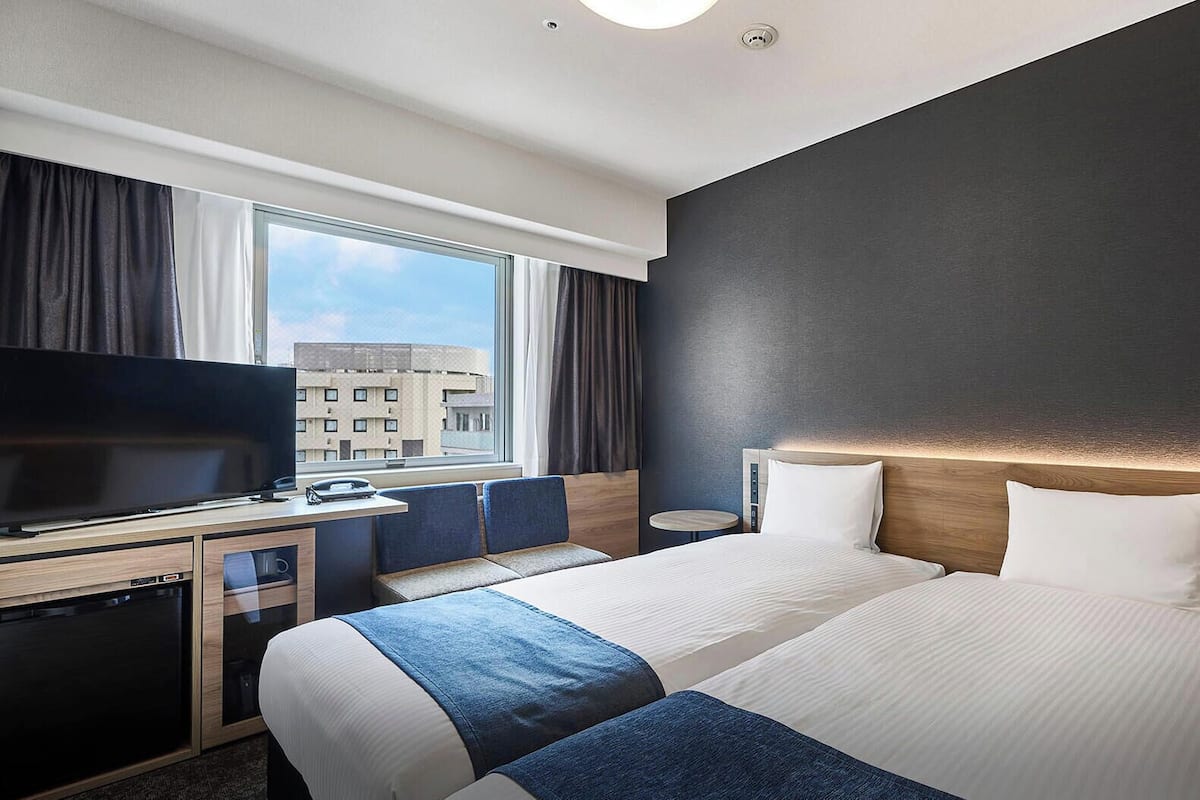 A hotel room with a TV, two beds, a desk, and a window with a city view.