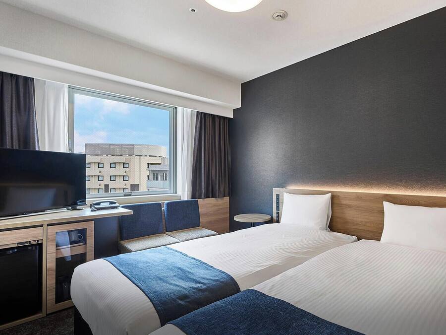 A hotel room with a TV, two beds, a desk, and a window with a city view.