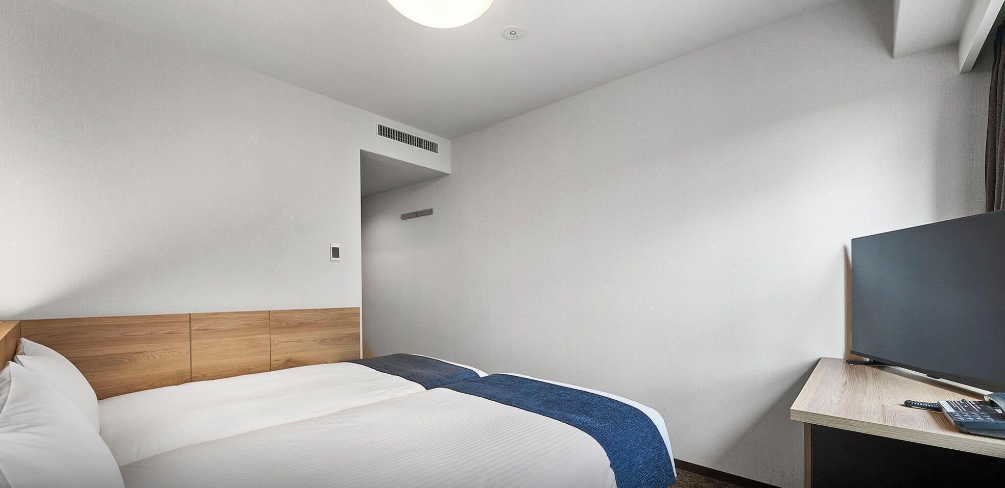 A hotel room with a bed, a TV, and a desk with a remote control.