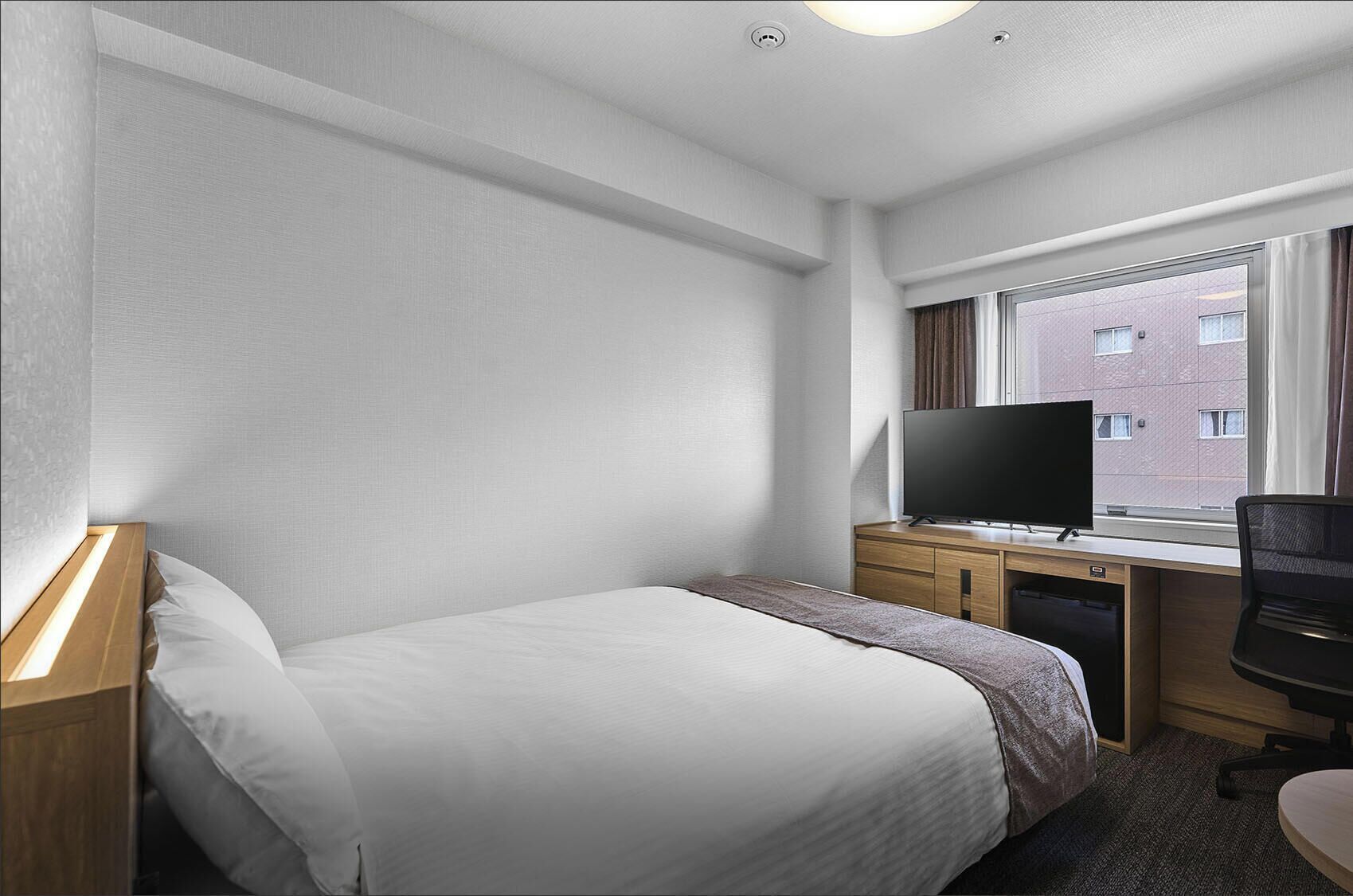 Une chambre d’hôtel avec un lit, un bureau avec une télévision, une chaise et une fenêtre donnant sur des immeubles.