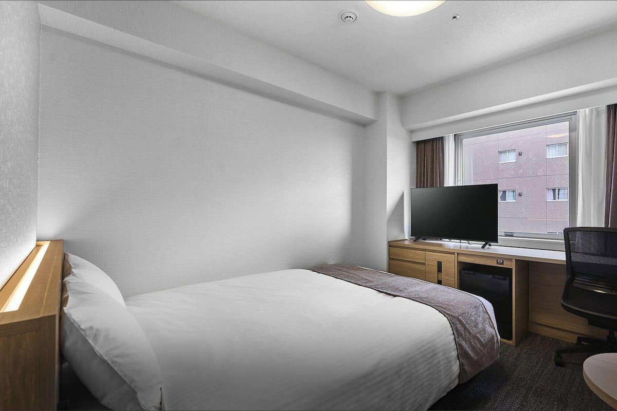 A hotel room with a bed, a desk with a TV, a chair, and a window with a view of buildings.