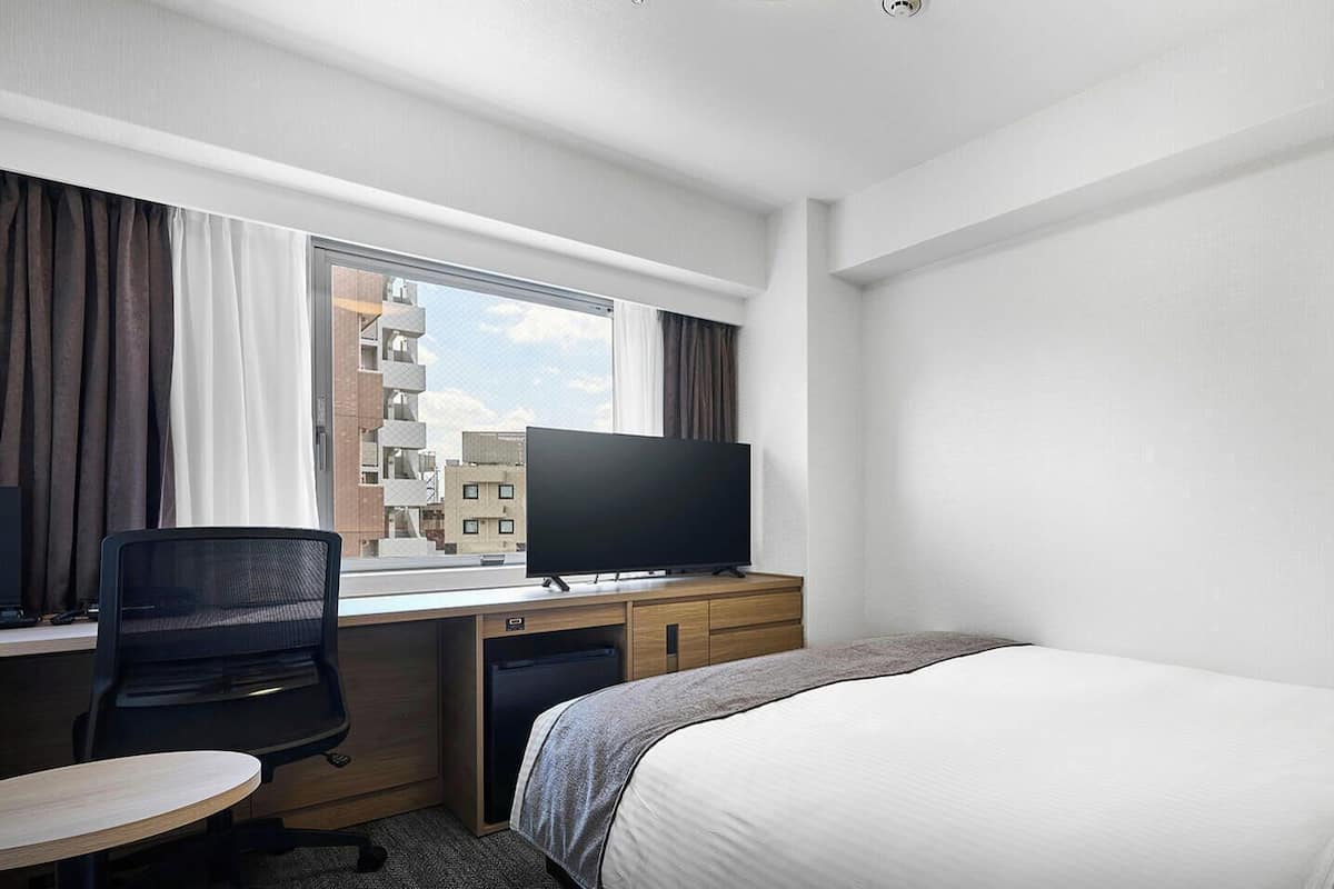 A hotel room with a bed, desk, chair, TV, and a window with curtains.