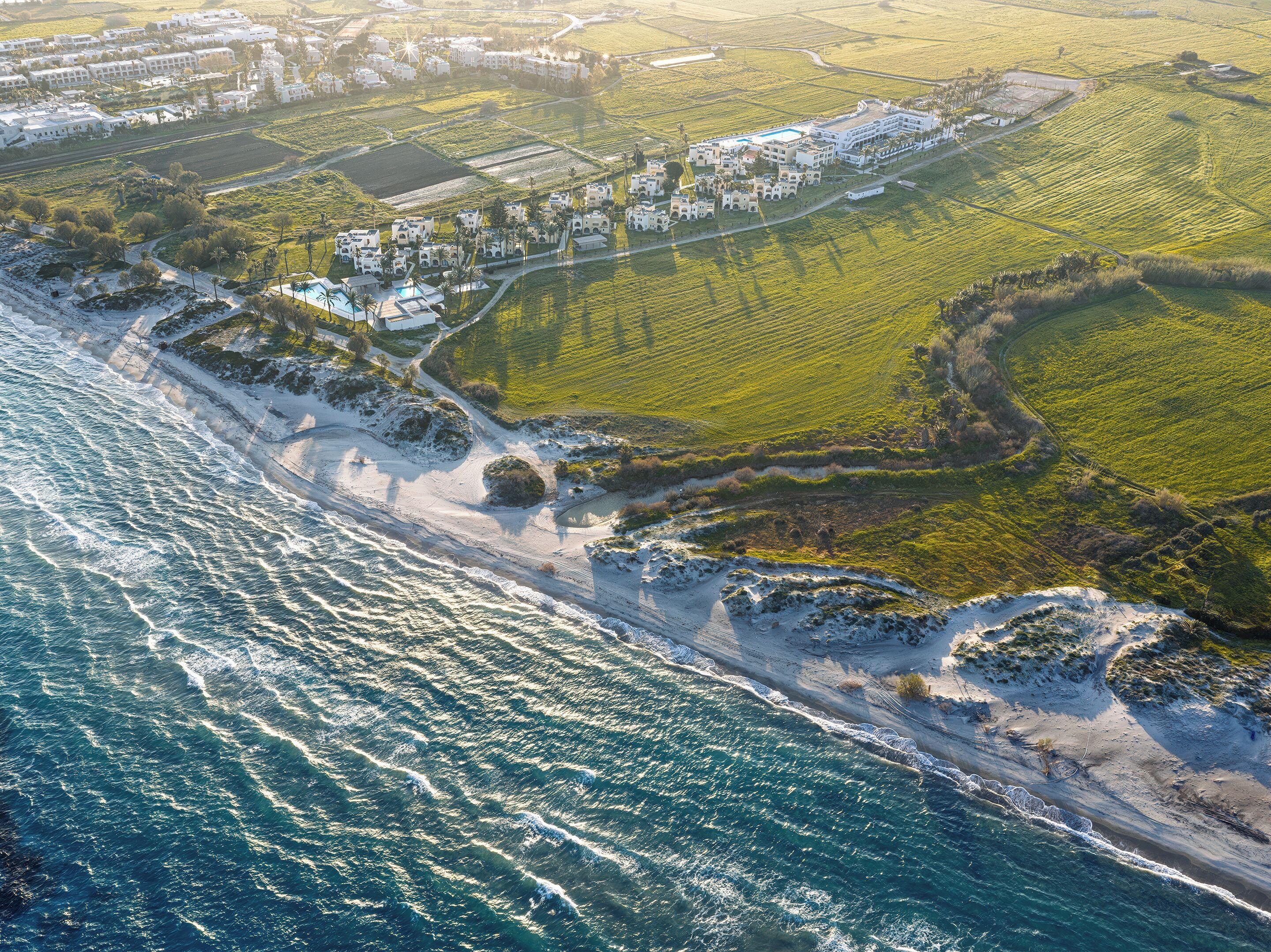 Vue aérienne d’une zone côtière comprenant un complexe hôtelier, des champs adjacents et une plage.