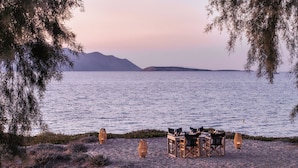 Disposición para comer frente al mar, con mesas y sillas, rodeada de vegetación natural y vista a una masa de agua tranquila con montañas a lo lejos.