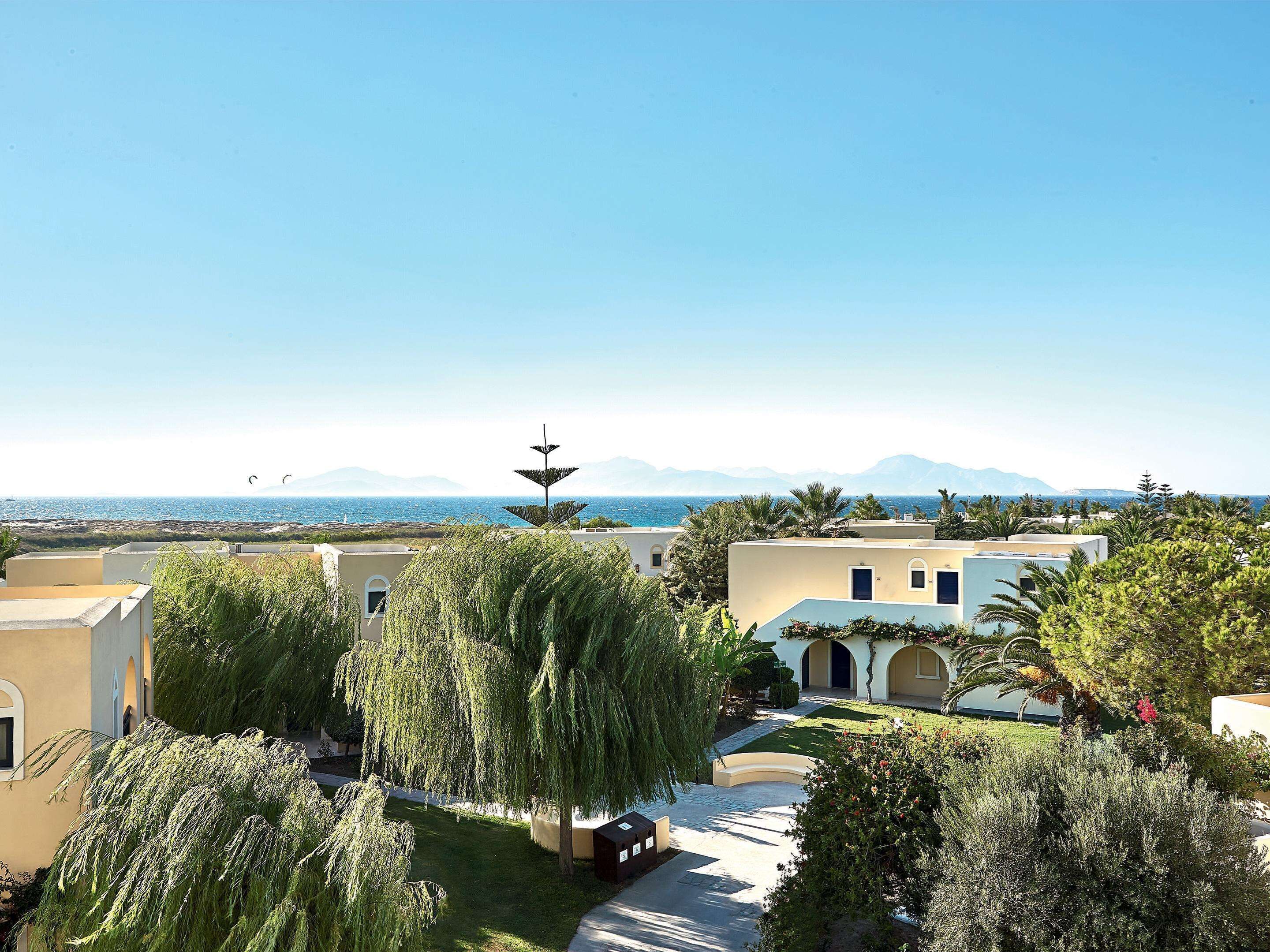 A coastal hotel with Mediterranean architecture, featuring terracotta walls, arched doorways, and a view of the sea and mountains.