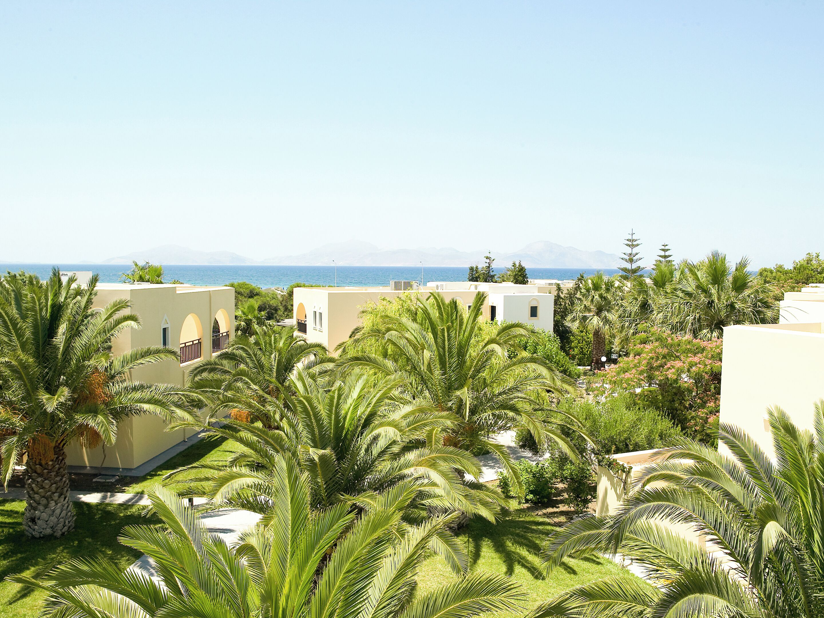 A coastal residential area with modern buildings, palm trees, and a clear view of the sea and mountains in the distance.