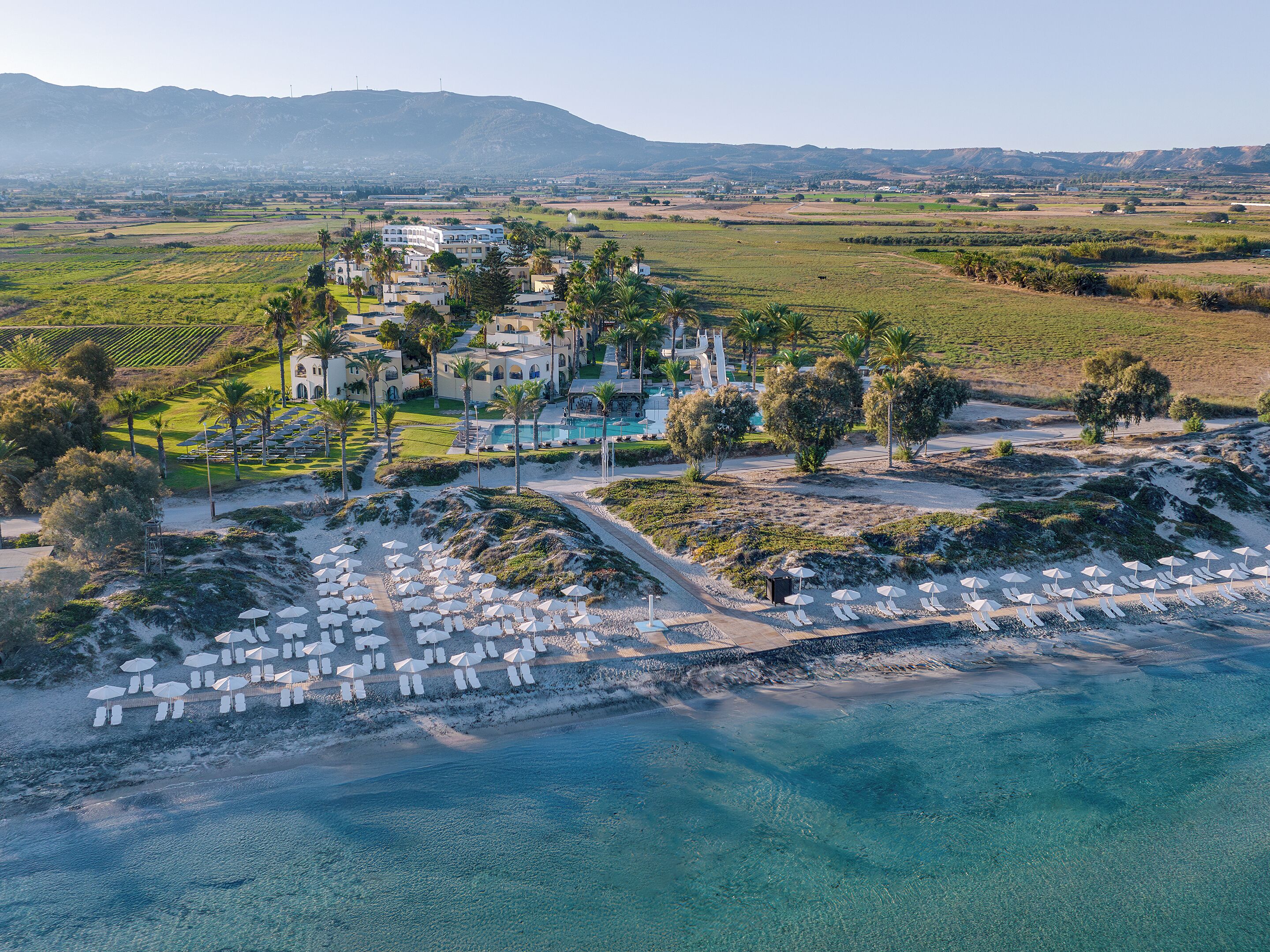 Vista aérea de un resort con piscina, sombrillas de playa y montañas al fondo.