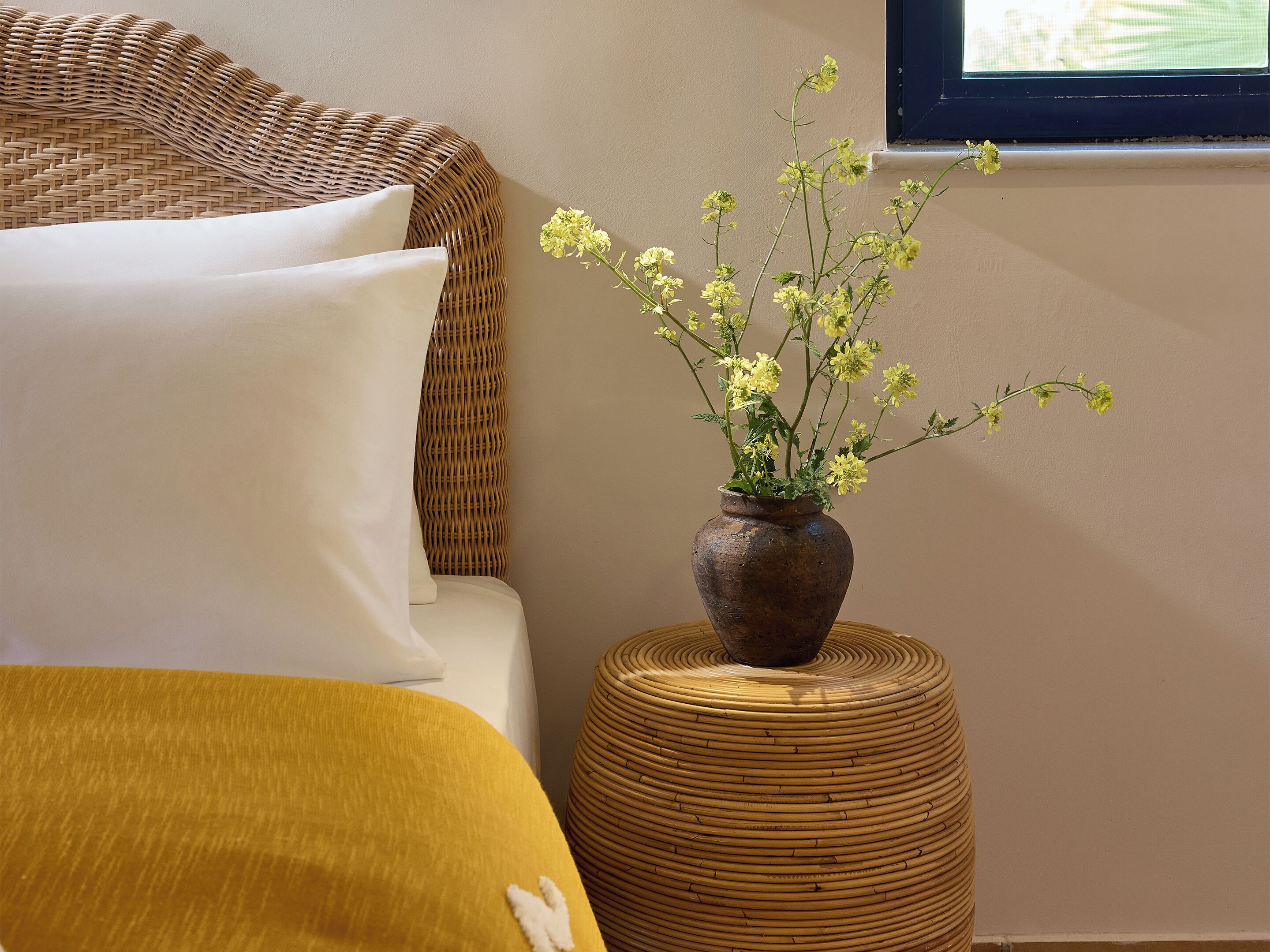 A bed with white and mustard pillows, a wicker headboard, a round woven stool, and a vase with yellow flowers on a wall.