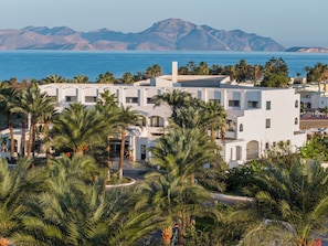 A white building with multiple balconies, surrounded by palm trees, with a view of the sea and mountains in the background. - Grecotel Casa Paradiso (Kos)