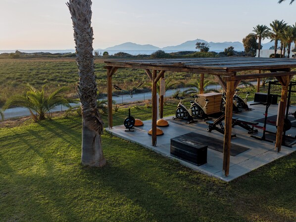 Un gimnasio al aire libre techado con equipamiento de ejercicio, un muelle de madera y vistas a un cuerpo de agua y montañas al fondo.