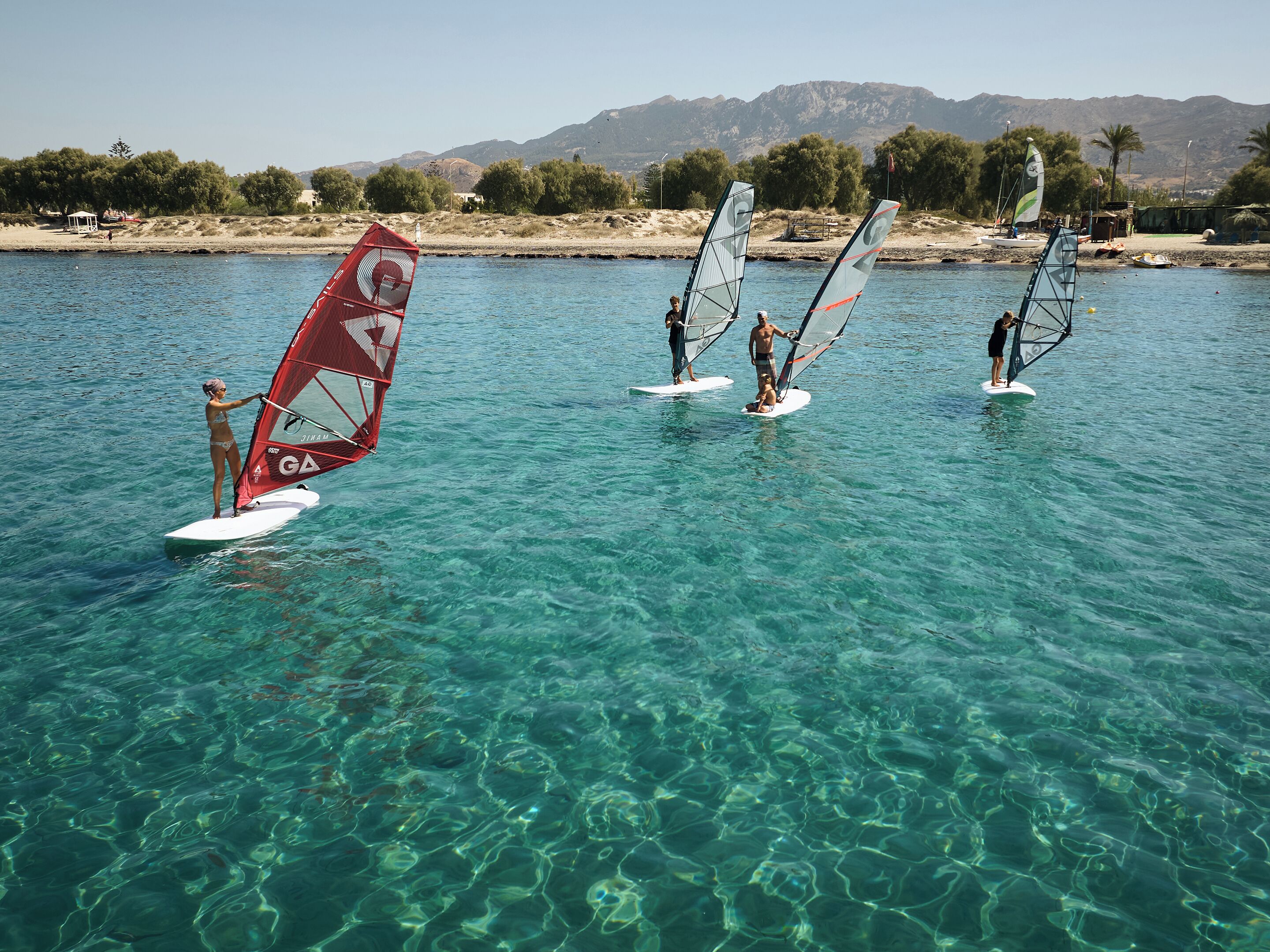 A group of people windsurfing on a clear, calm body of water with mountains in the background.