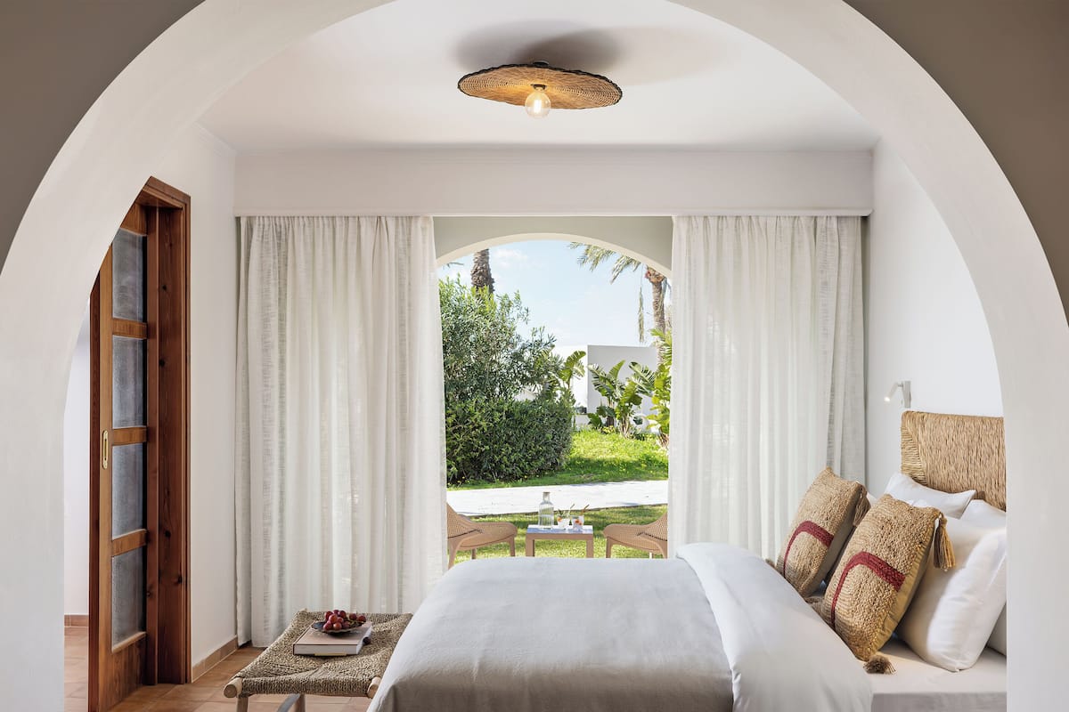 A bedroom with a bed, a small table with a book and fruit, a wicker chair, and a view of a garden through a window.