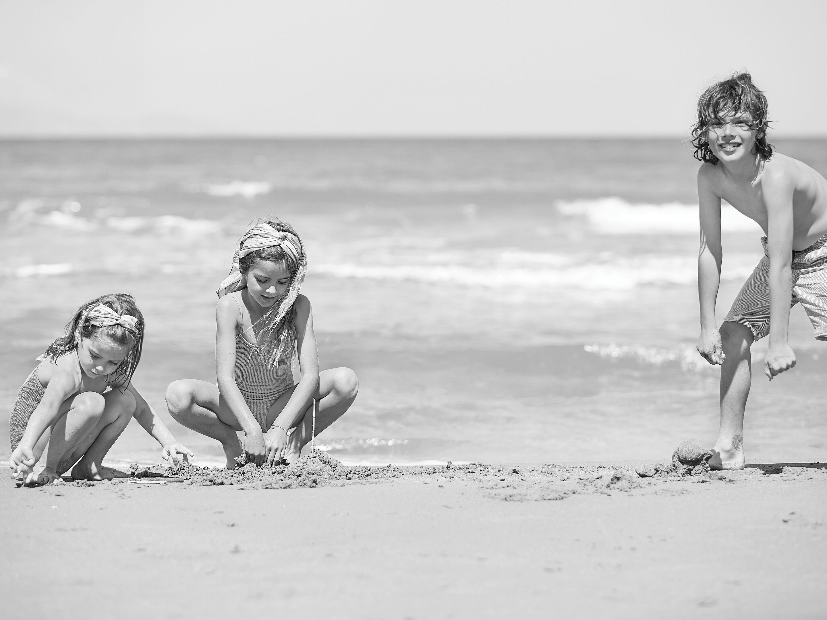 Trois enfants jouent sur une plage de sable, avec l’océan en arrière-plan.