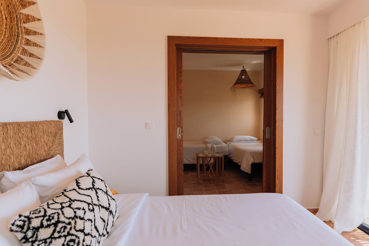 A bedroom with a large bed, a bedside lamp, and a view into an adjacent room with a table and chairs.