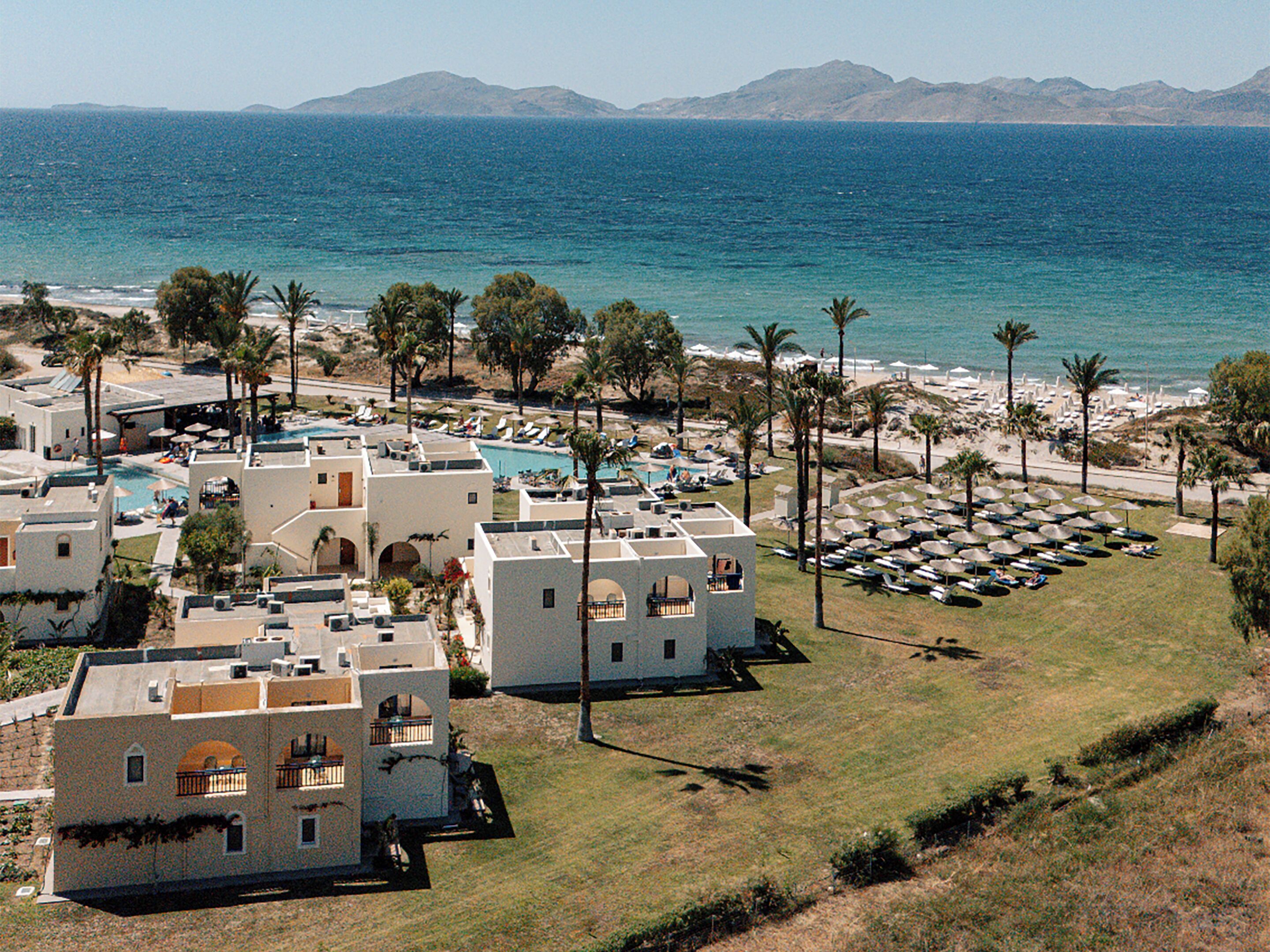A coastal resort with multiple buildings, a swimming pool, and a beach area with umbrellas.