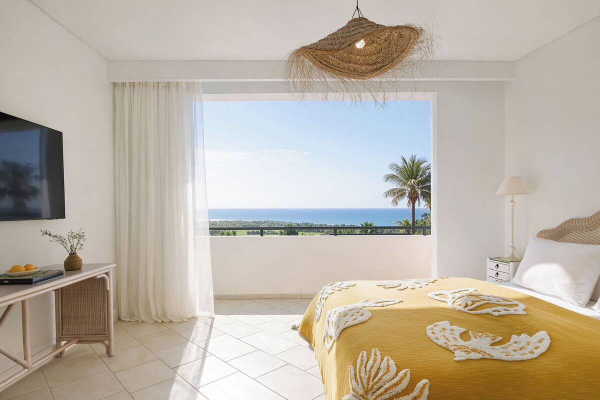 A bedroom with a bed, a TV, a desk, and a view of the ocean.